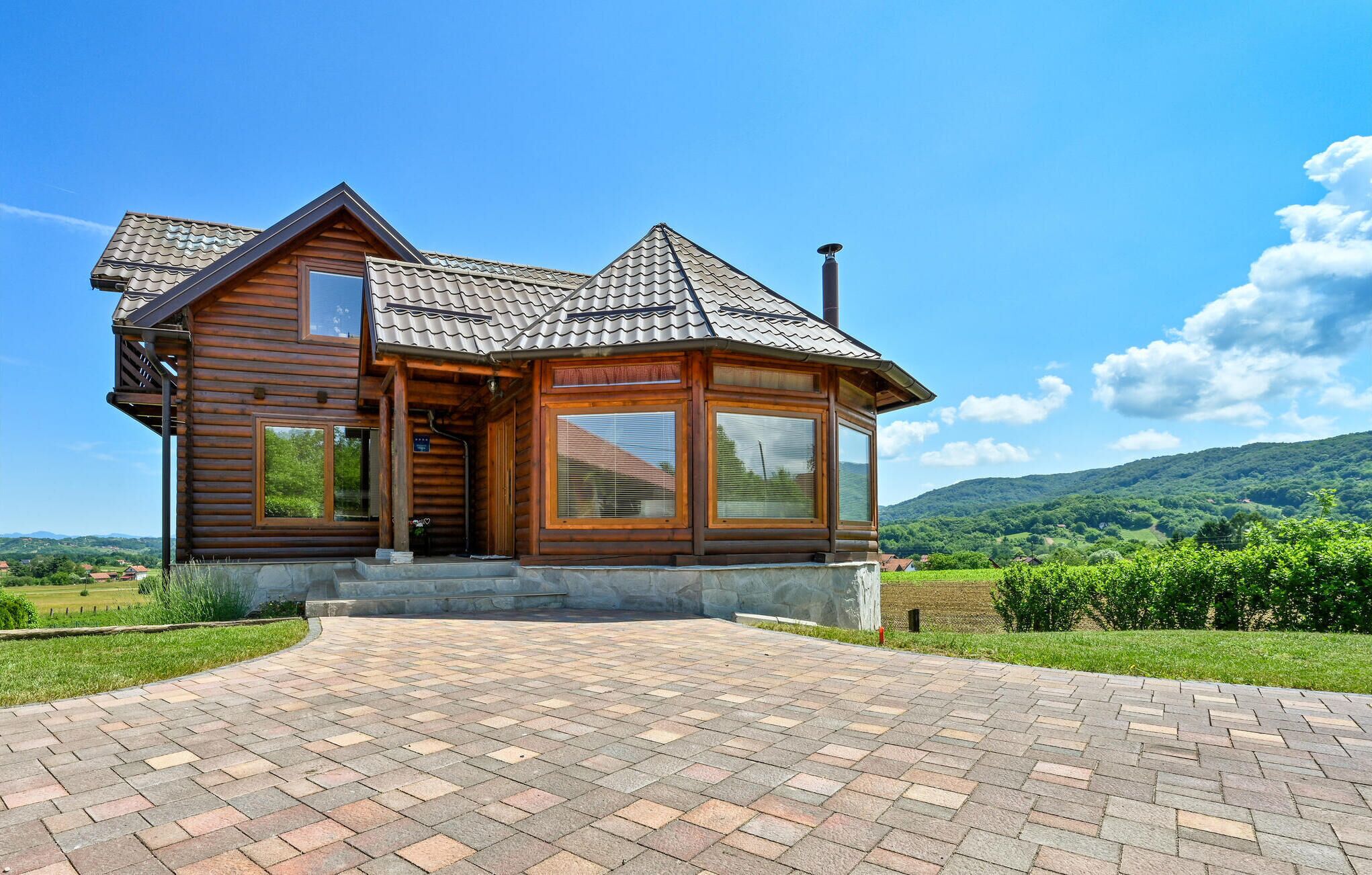 A wooden cabin with a tiled roof, large windows, and a paved driveway leading to the entrance.