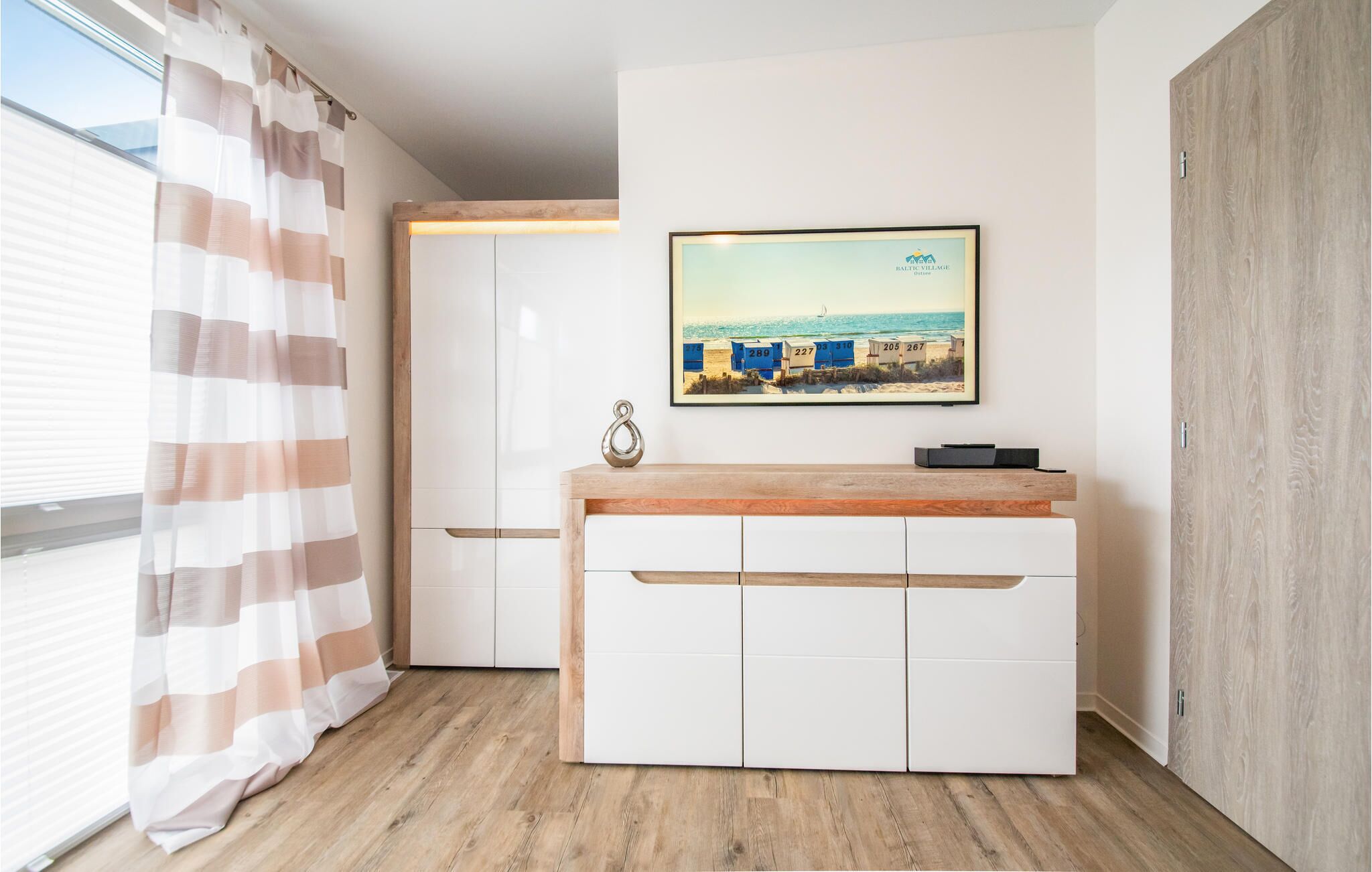 Ein modernes Zimmer mit Holzboden, weißen Schränken, einem gerahmten Bild mit Strandmotiv und einem Streifenmuster-Vorhang.