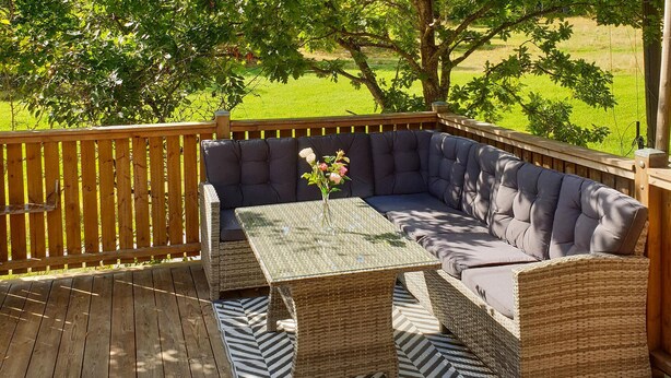 A deck with a wicker sofa and a table, surrounded by a wooden deck and a grassy field.