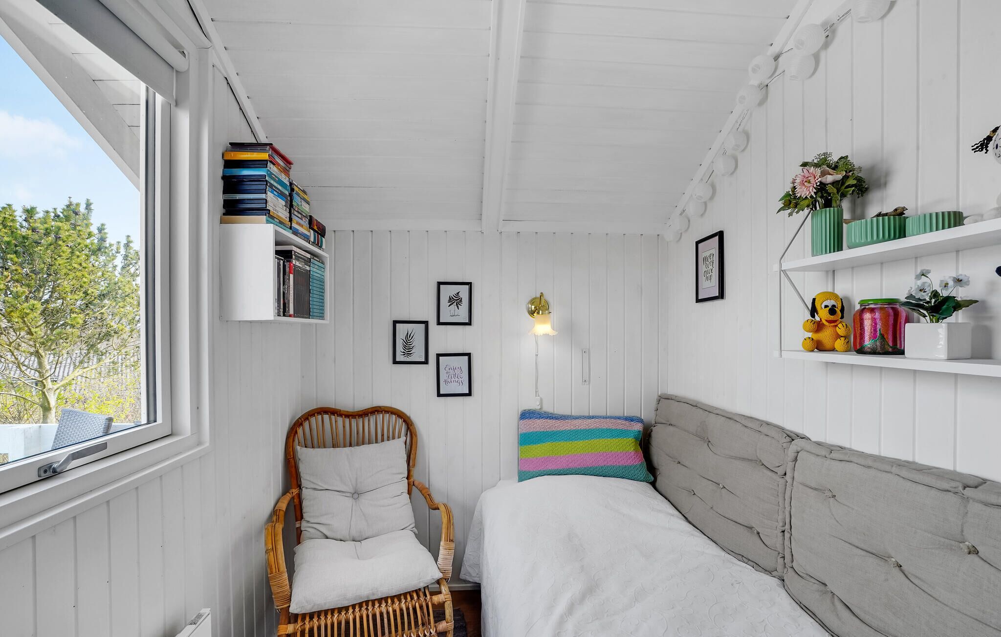 Una habitación con una cama, una silla de mimbre, una estantería y un ventanal con vistas a árboles.