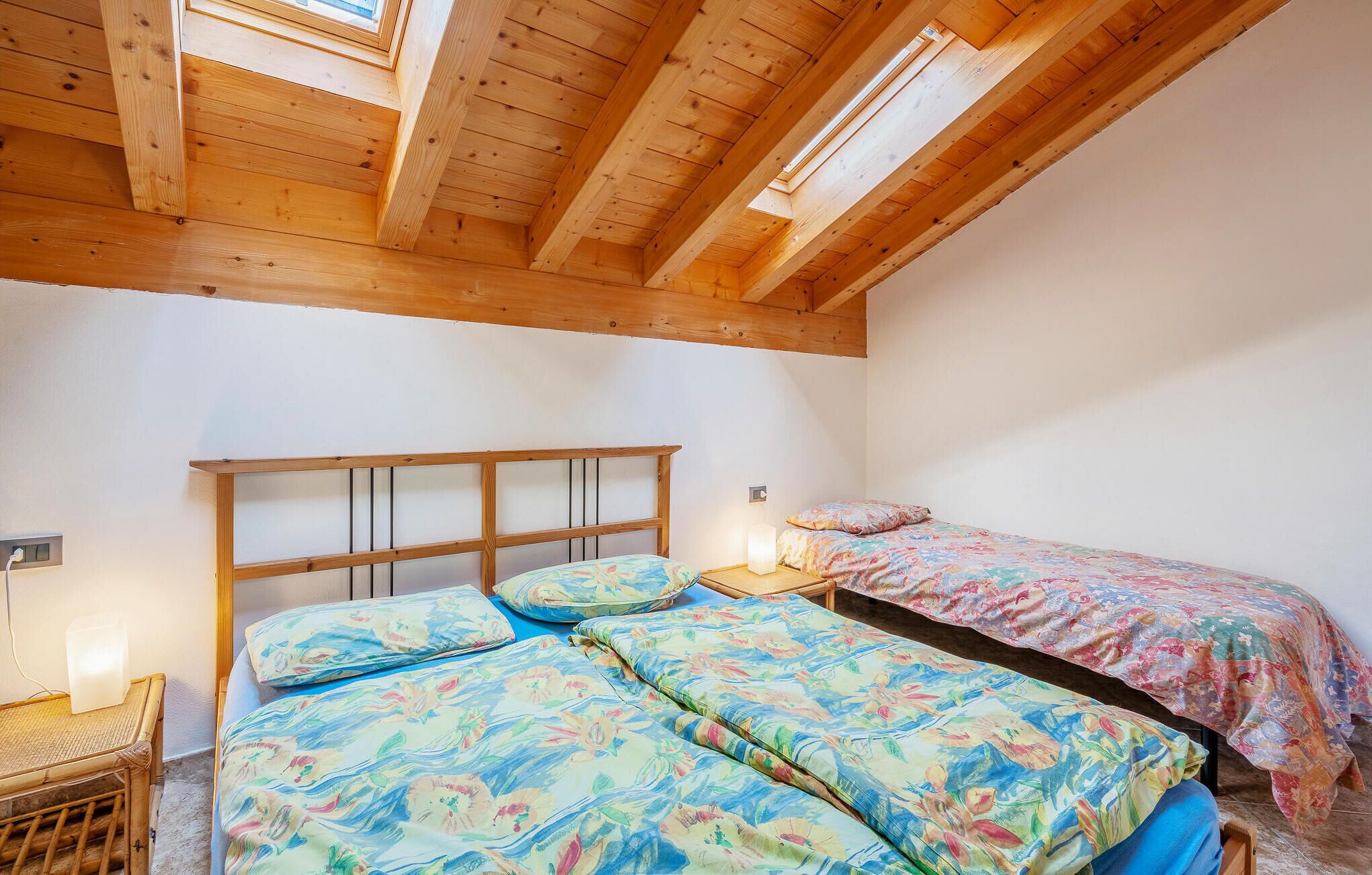 A bedroom with two beds, wooden beams, a skylight, and a bedside table with a lamp.