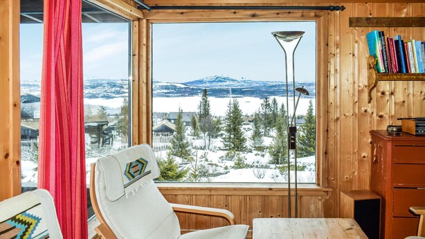A cozy cabin room with a large window offering a snowy mountain view, a wooden chair, a table, and a bookshelf with books. - 2 bedroom beautiful home in Gol (Gol)