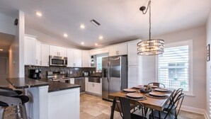 Una cocina moderna con armarios blancos, electrodomésticos de acero inoxidable, una mesa de comedor de madera y una lámpara colgante.