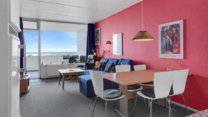 Un salón moderno con una pared de acento roja, un sofá azul, una mesa de comedor de madera y vistas al océano.