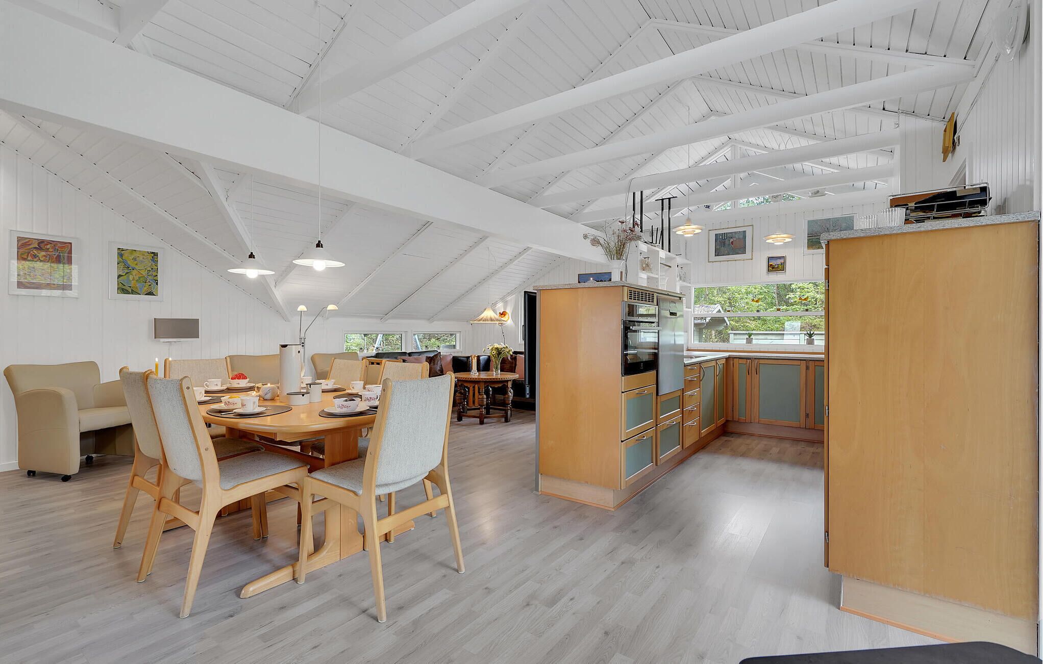 Un menjador ampli amb un sostre alt i bigues de fusta, una cuina amb armaris de fusta, i vistes a zones verdes a l'exterior.