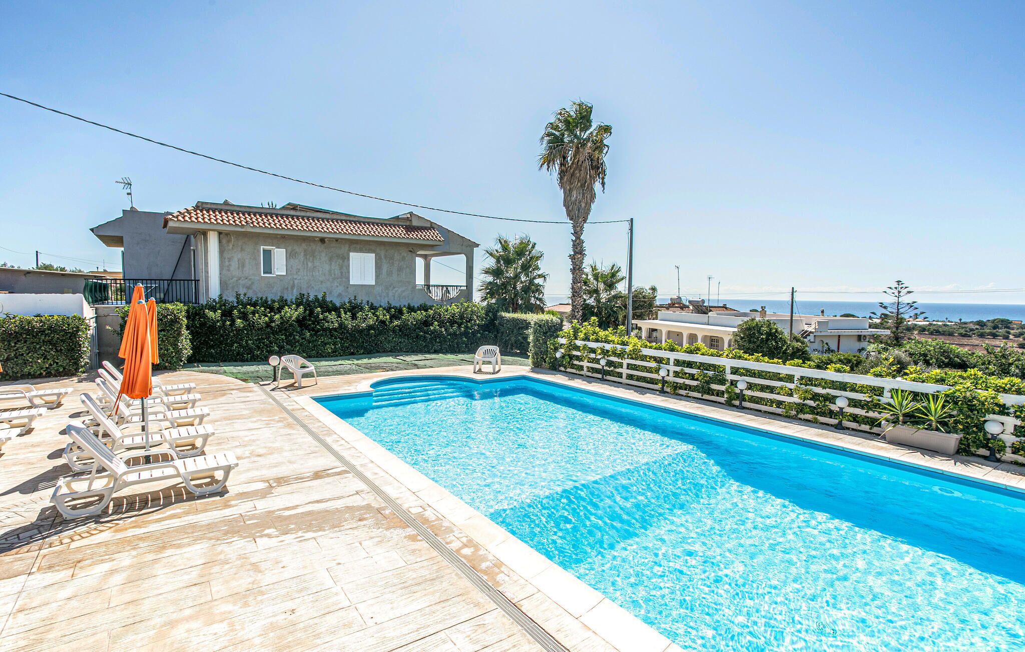 A swimming pool with lounge chairs and umbrellas, surrounded by a white fence and palm trees, with a view of the sea and a building in the background.