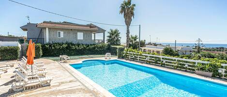 A swimming pool with lounge chairs and umbrellas, surrounded by a white fence and palm trees, with a view of the sea and a building in the background.