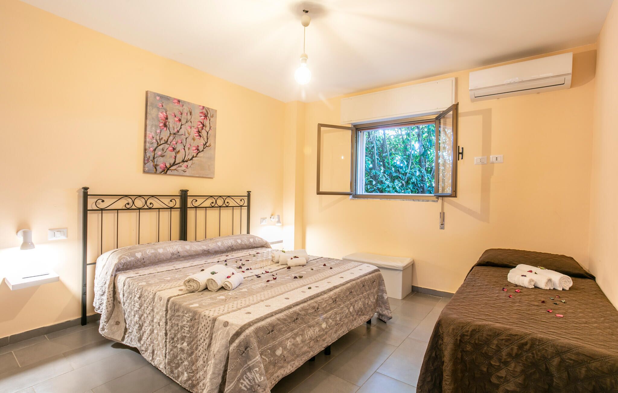 A bedroom with a bed, a bedside table, a window with a view of greenery, and an air conditioning unit.