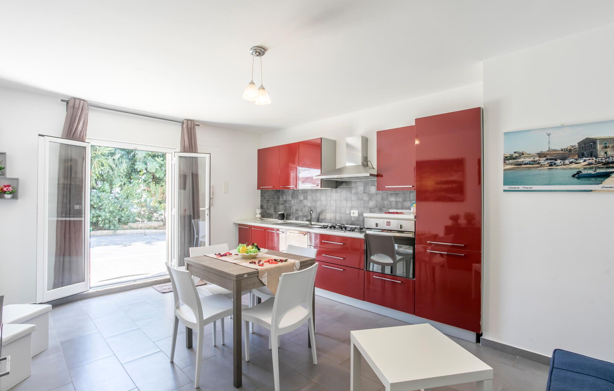 A modern kitchen with red cabinets, a dining table with chairs, and a view of the sea through the window.