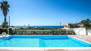 A swimming pool with a view of the sea, surrounded by greenery and a white fence.