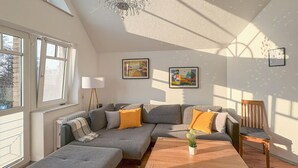 Una sala moderna con un sofá seccional gris, una mesa de centro de madera y una lámpara de pie. Hay dos ventanas que aportan luz natural, y se exhiben obras de arte en las paredes.