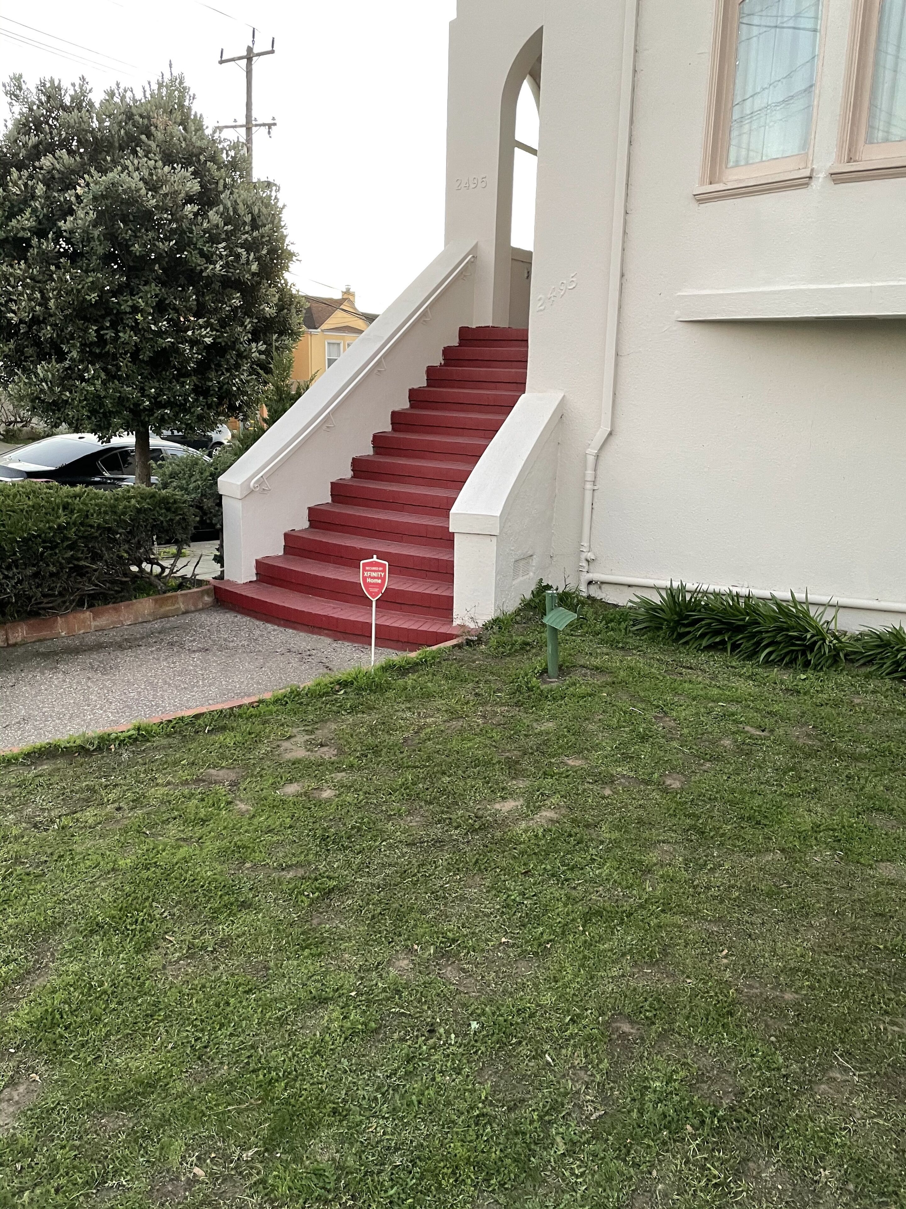 A residential building with a red staircase leading to a white door, a small garden area, and a parked car.
