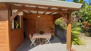 Une pergola en bois avec une table et des chaises, entourée de plantes en pot et d’un jardin.