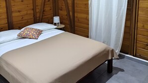 Un lit simple avec une couvre-lit beige, un oreiller blanc et une petite table de chevet avec une lampe. Un rideau blanc est accroché à côté du lit, et un ventilateur de plafond est fixé au mur.