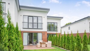 Ein modernes, zweistöckiges Haus mit Terrasse, einer Glastür und einer kleinen Sitzecke mit Korbmöbeln.