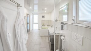 Ein modernes Badezimmer mit einer großen Badewanne, einem Waschbecken und einer Toilette. Links befindet sich ein Handtuchhalter und über dem Waschbecken ein Spiegel.