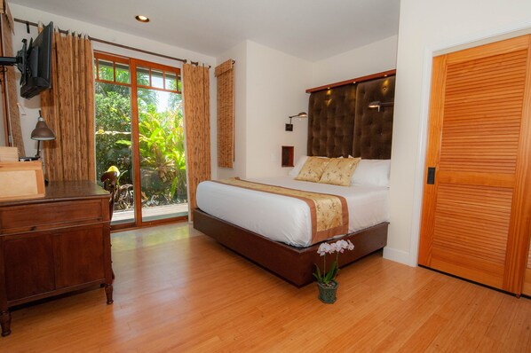 A bedroom with a large bed, wooden floor, a desk, a TV, and a sliding glass door to a garden.