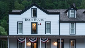 A two-story building named "Bush House" with a porch featuring white wicker chairs and a flower bed. - Bush House Inn (Index)