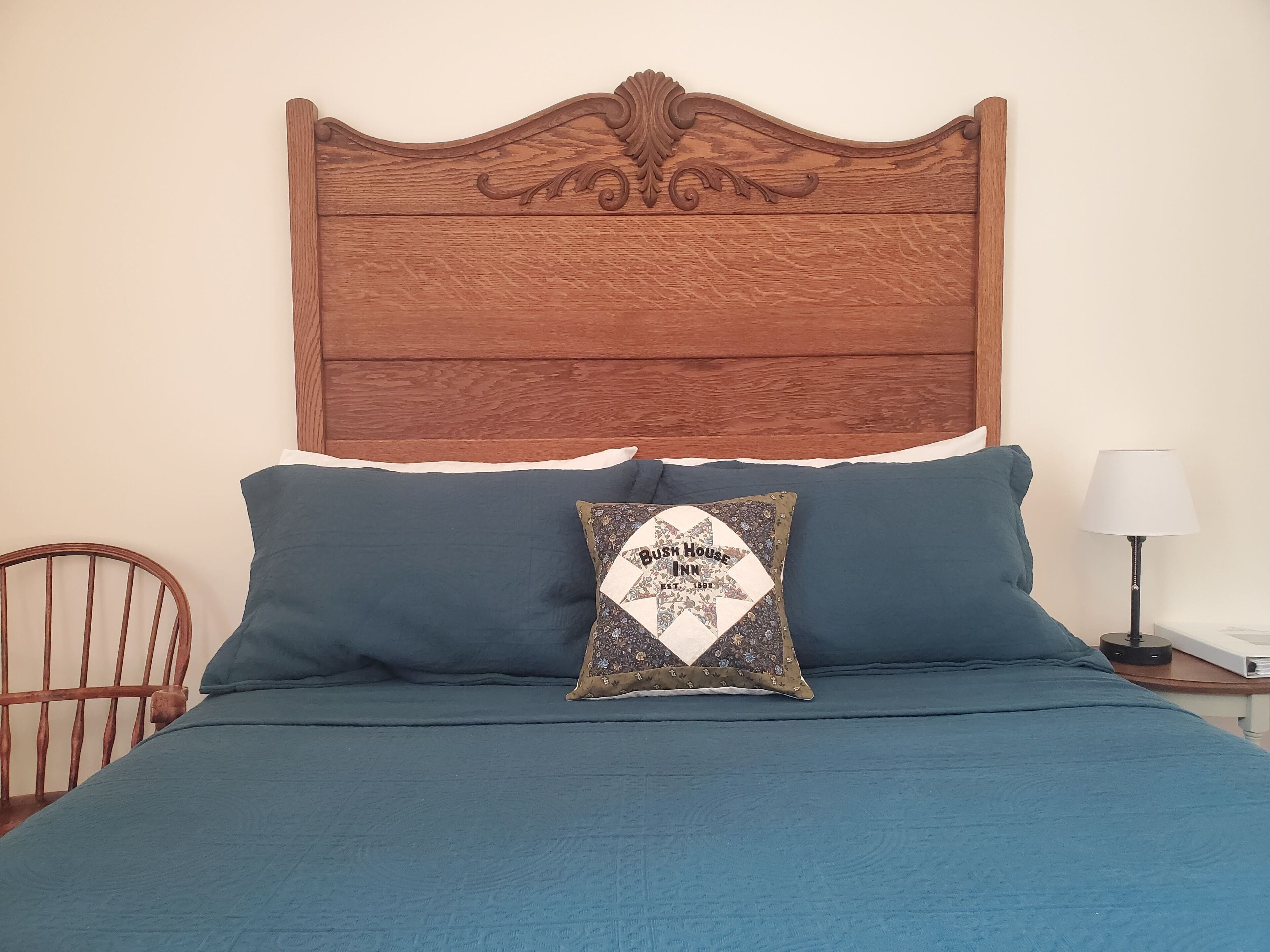 Una cama con cabecera de madera, dos almohadas, un cojín decorativo y una mesita de noche con una lámpara.