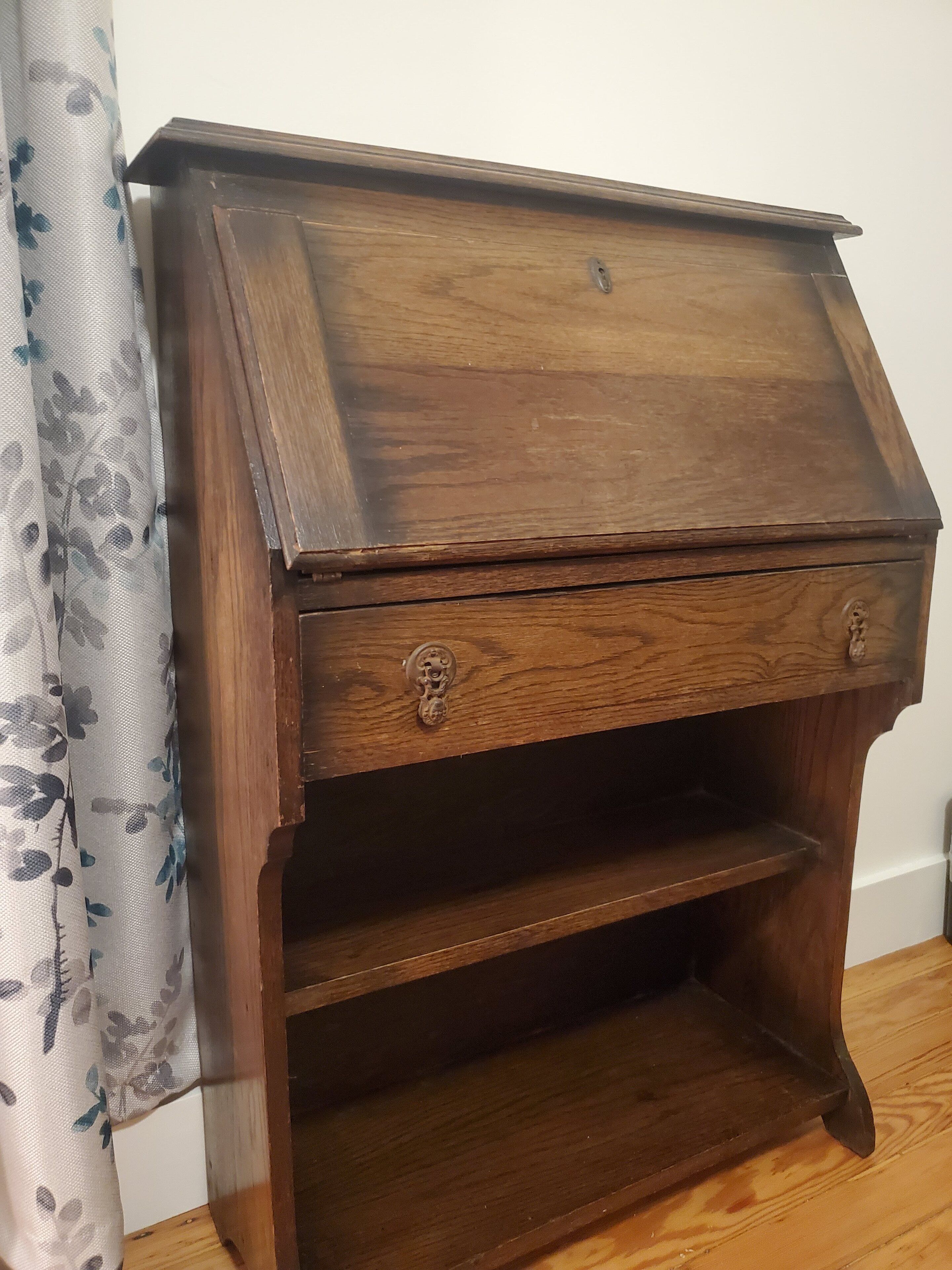 A wooden side table with a drawer and a shelf, positioned against a wall with a floral curtain.