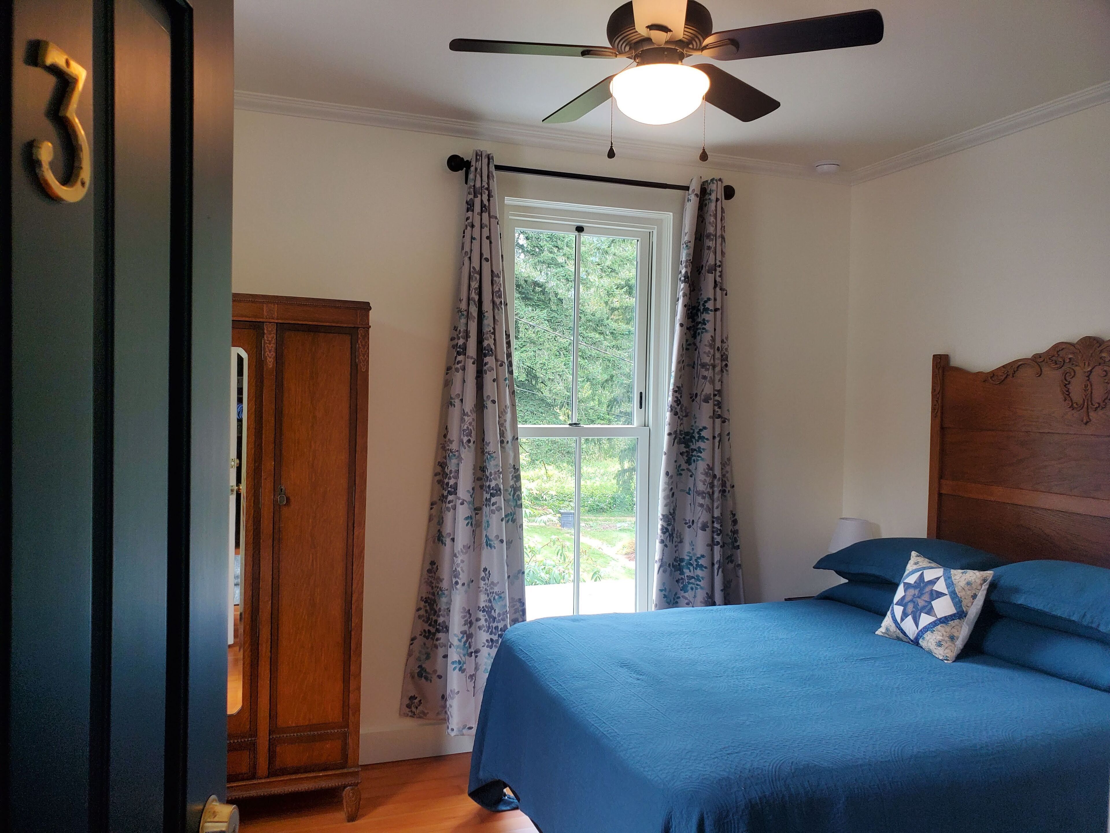 A bedroom with a bed, a ceiling fan, a window with curtains, and a wooden wardrobe.