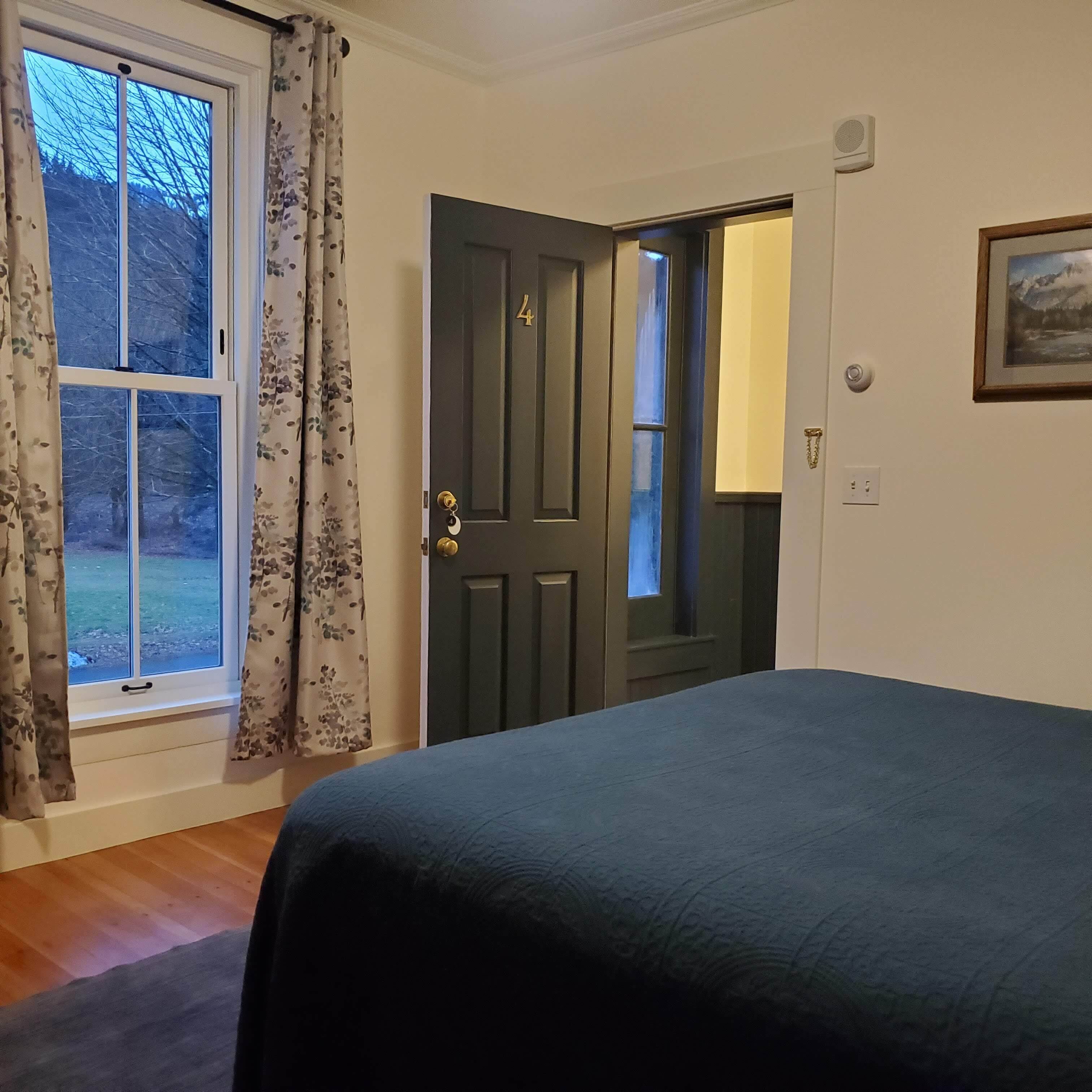 A bedroom with a bed, a door, a window with curtains, and a framed picture on the wall.
