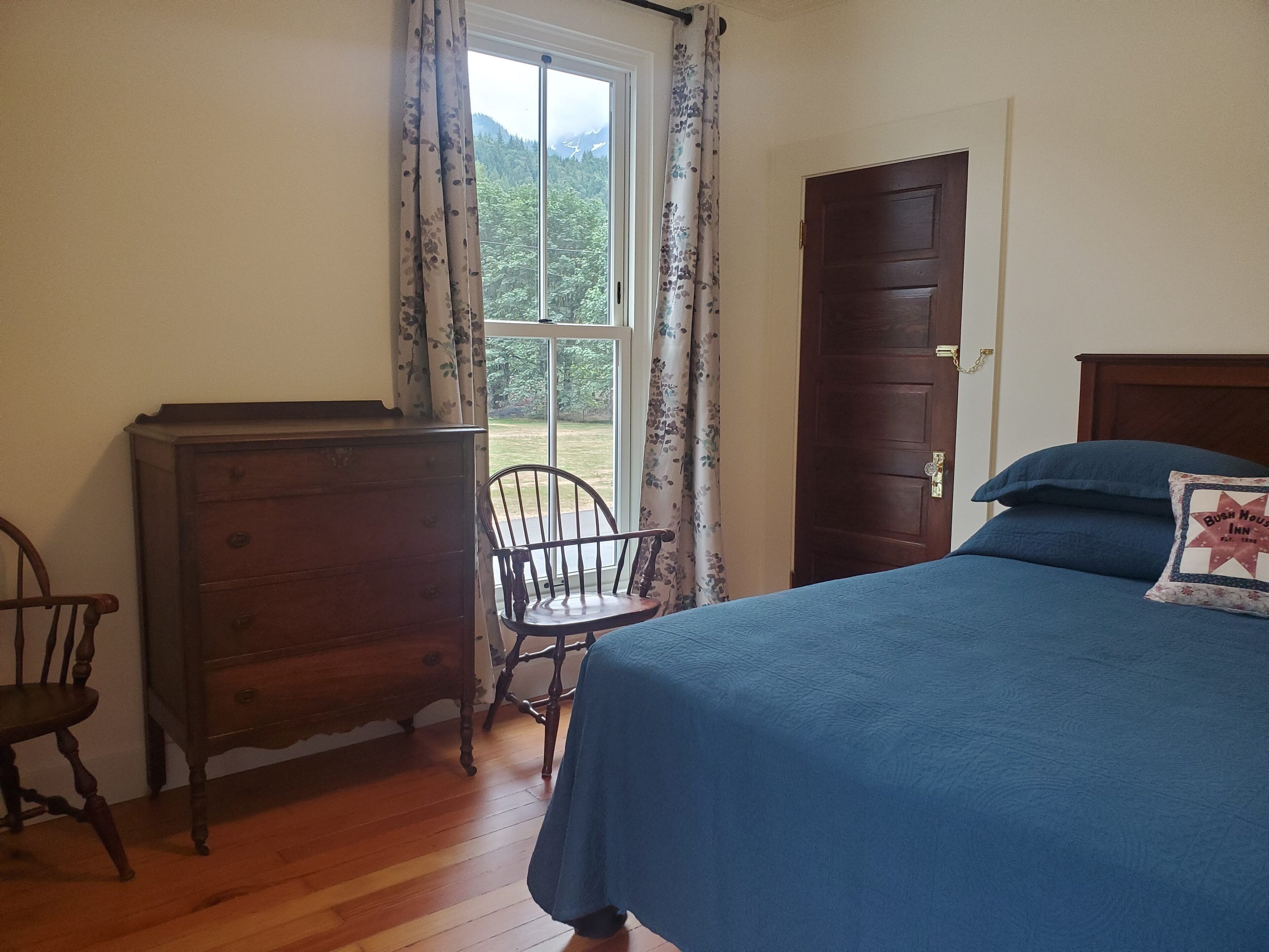 Ein Schlafzimmer mit einem hölzernen Schminktisch, einem Stuhl, einem Bett mit blauer Bettdecke und einer Tür, die auf einen Balkon mit Blick auf Grünflächen und Berge führt.
