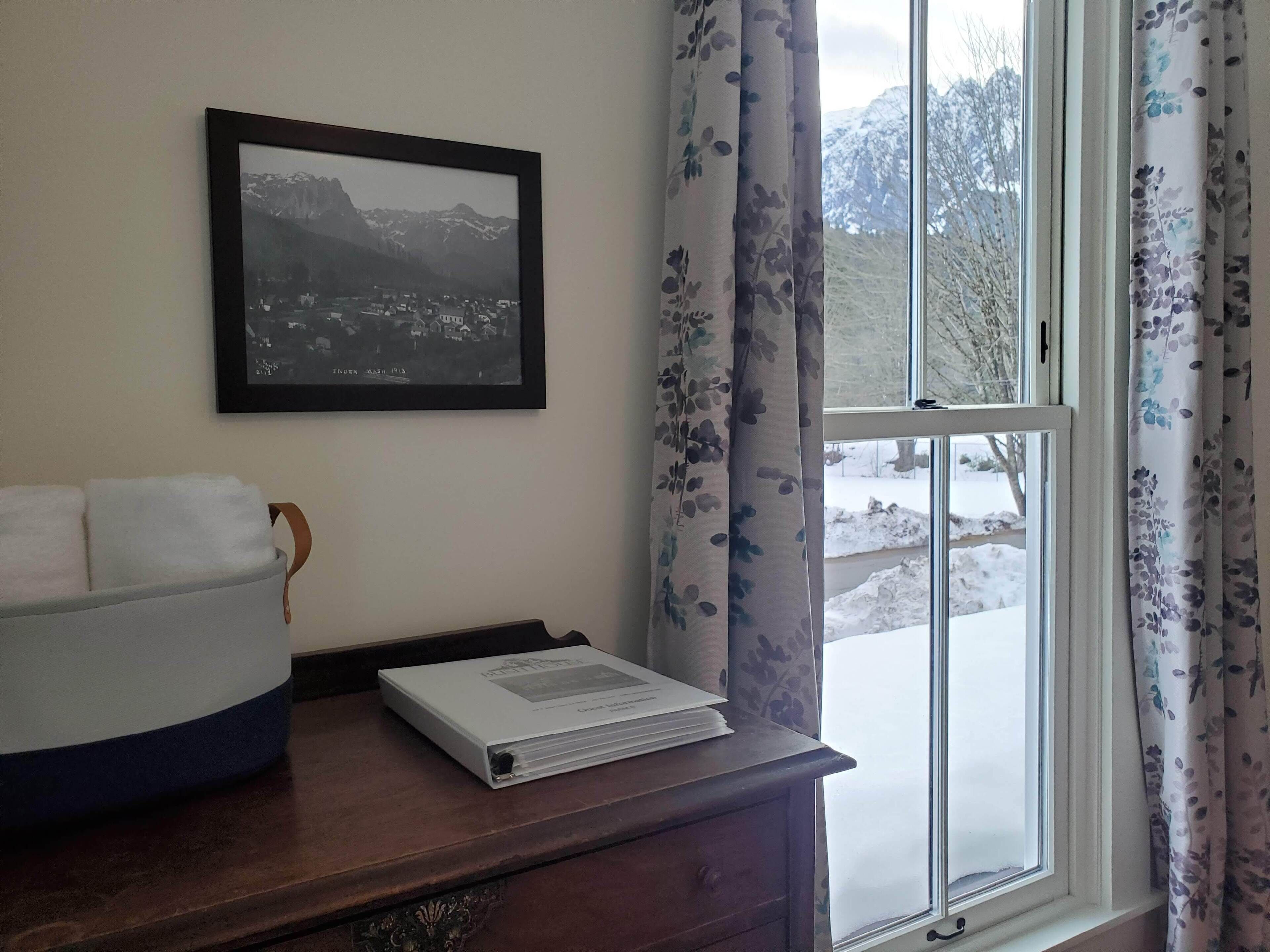 A room with a framed black and white photograph on the wall, a window with floral curtains, and a wooden desk with a stack of papers and a white container.