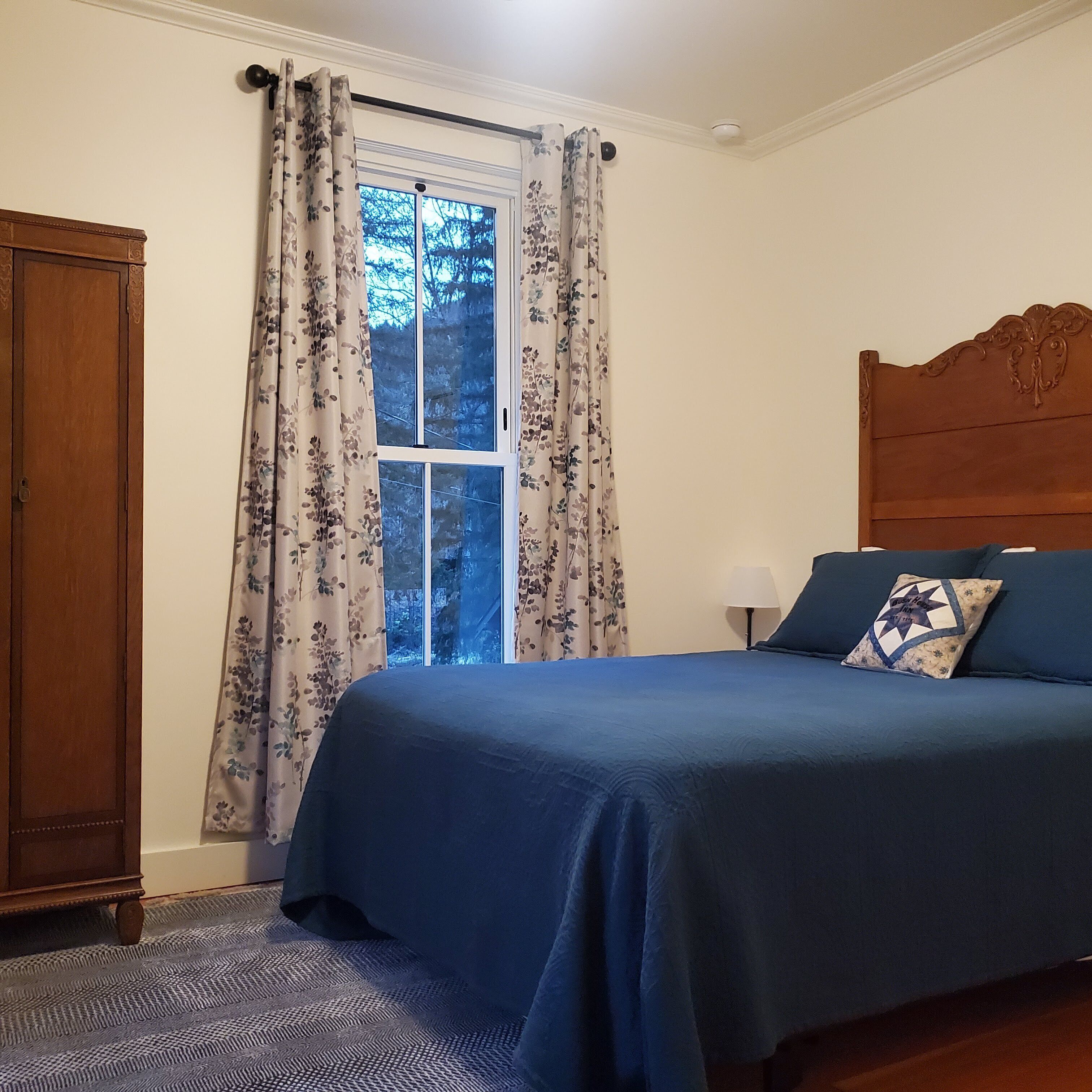 Un dormitorio con cama, cabecera de madera, ventana con cortinas y mesita de noche con lámpara.