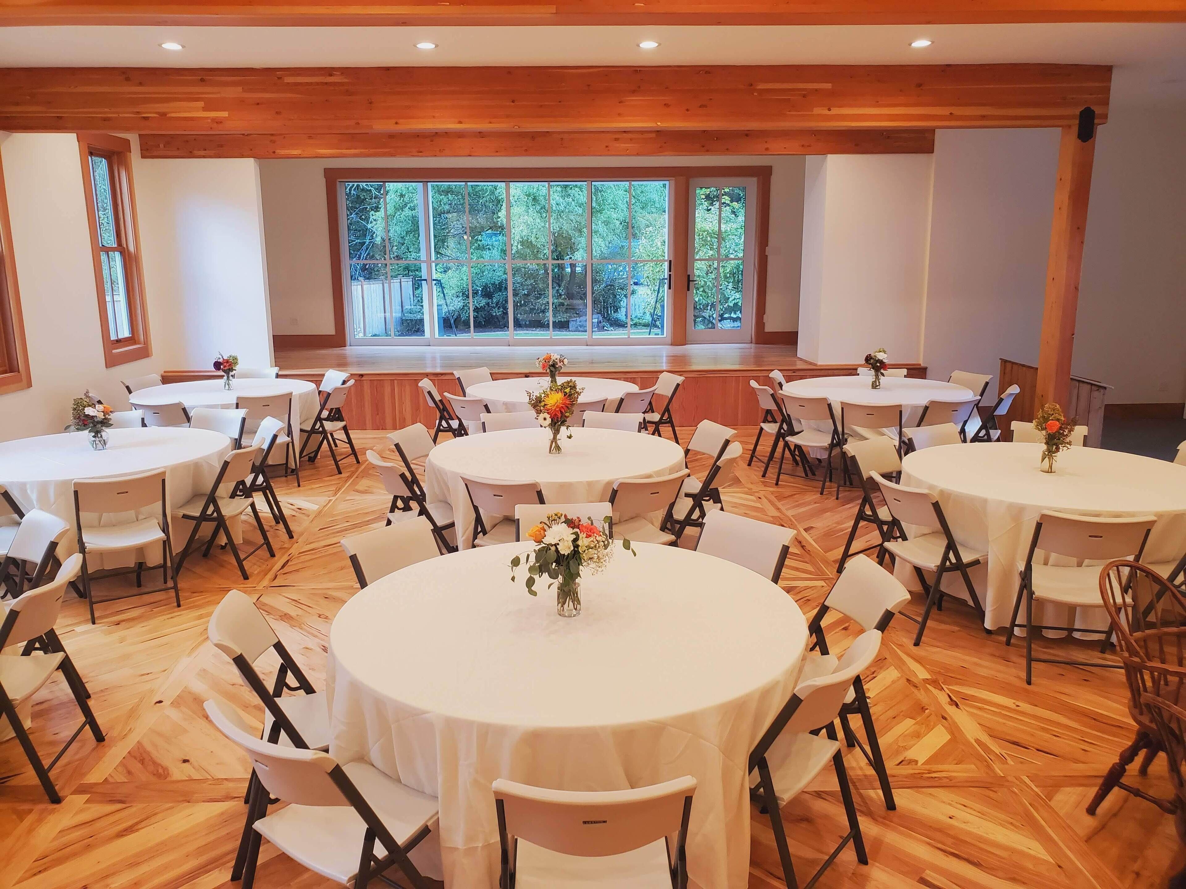 Un salón de conferencias con varias mesas redondas, sillas blancas y centros de mesa con flores.
