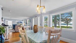 A dining area with a large table, chairs, and a chandelier. There are windows with a view of trees and a body of water.