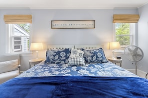 A bedroom with a bed, two bedside tables, a wall-mounted fan, and a window with a view of trees.