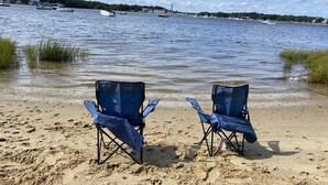 Two blue folding chairs on a sandy beach with a view of a lake and boats in the distance.