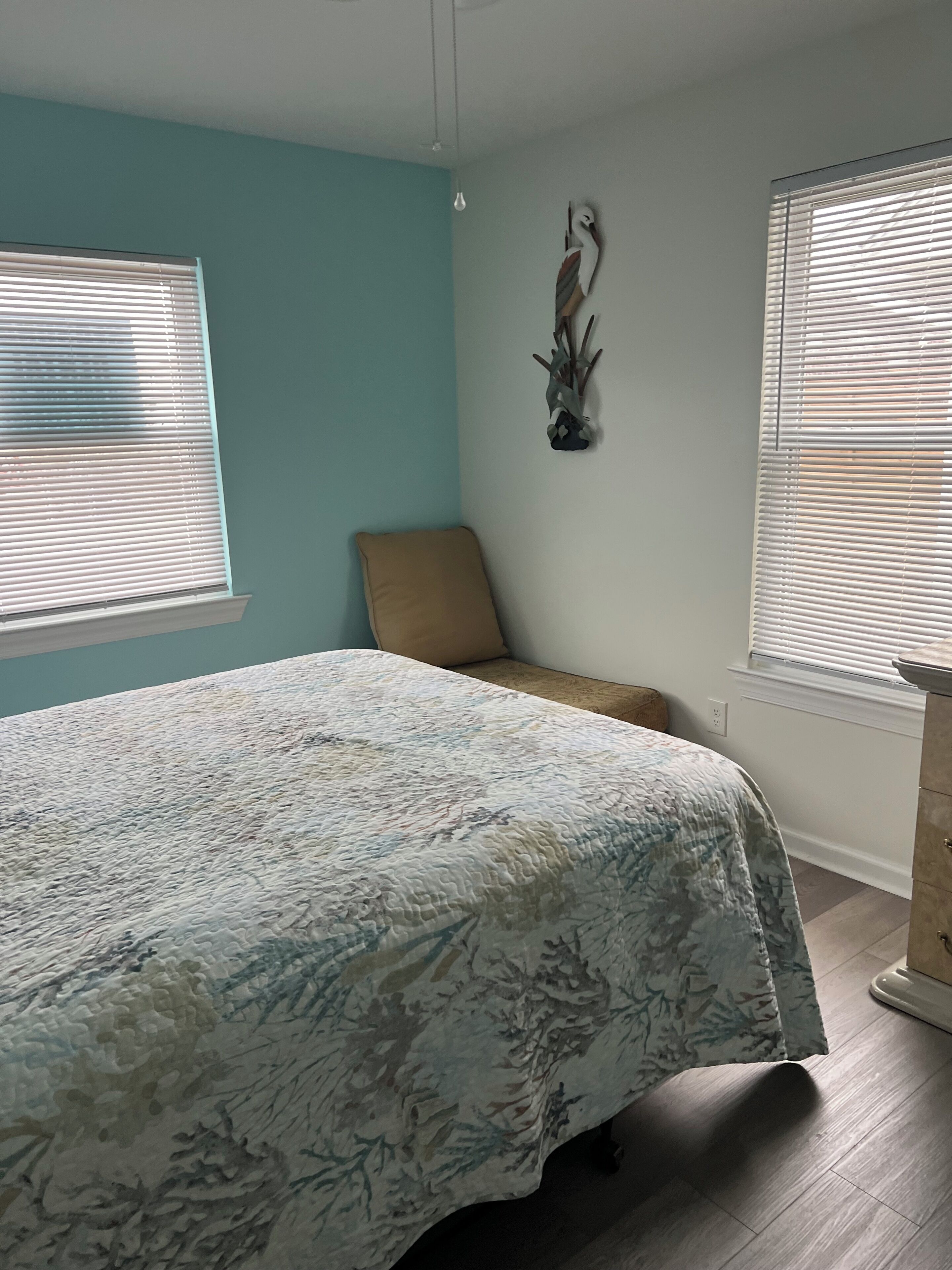 A bedroom with a bed, a chair, a wall-mounted decoration, and two windows with blinds.