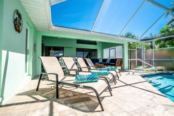 A covered outdoor area with lounge chairs, a pool, and a patio with a table and chairs.