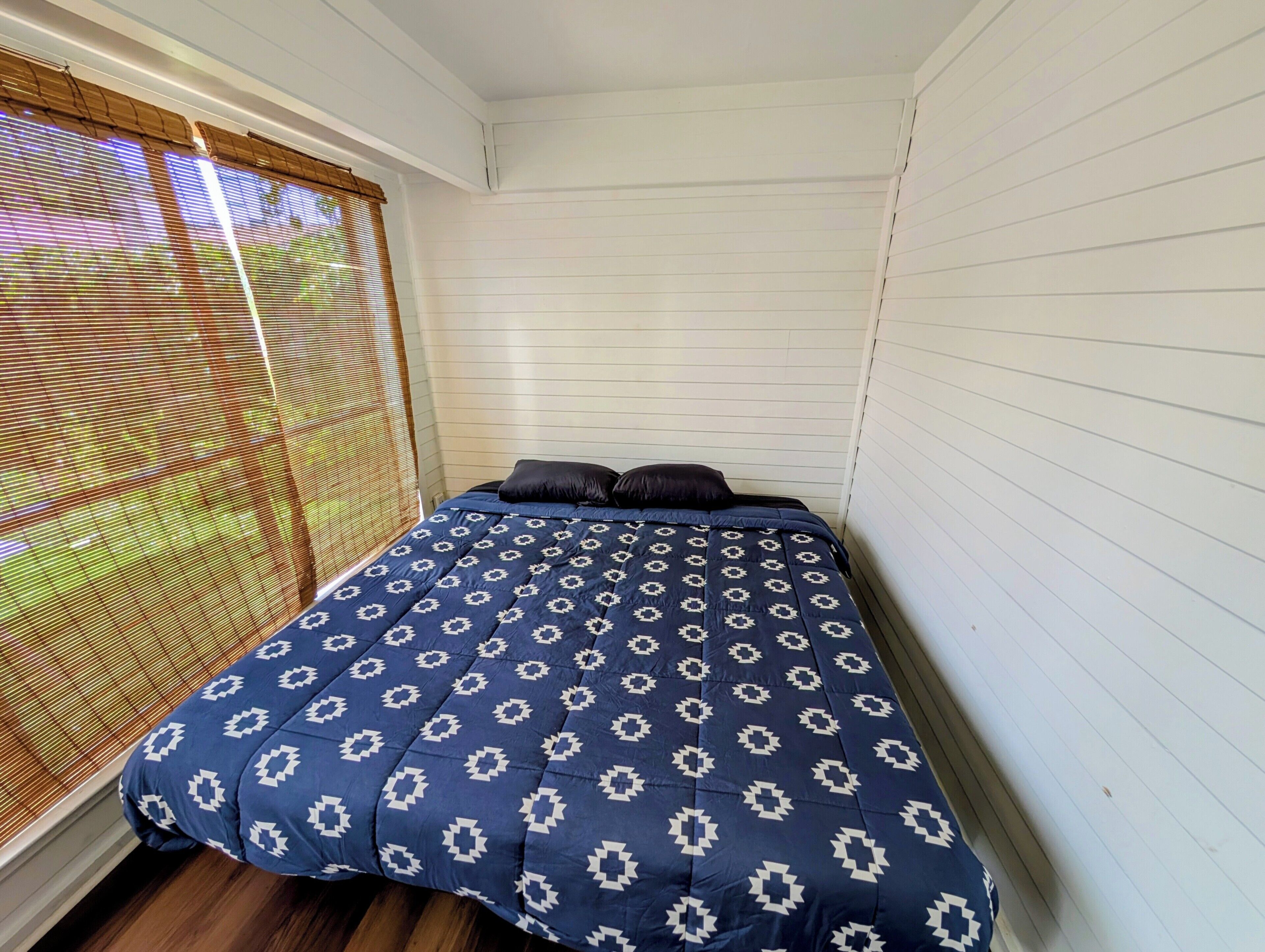 Una habitación pequeña con cama individual, colcha estampada, ventana con persianas y piso de madera.