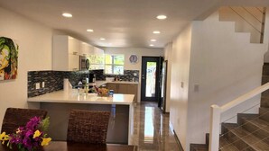 Una cocina moderna con una isla central, una zona de comedor con sillas de mimbre y una escalera que sube a un nivel superior.