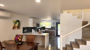 Una cocina moderna con una isla central, un comedor con sillas de mimbre y una escalera que sube a un nivel superior.