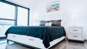 Un dormitorio moderno con un ventanal grande, una cama con colcha verde azulada, una mesita de noche con lámpara y un cuadro en la pared.