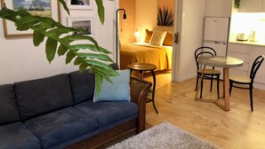 Ein Wohnzimmer mit einer Couch, einem kleinen runden Tisch und Stühlen. Ein Bett mit gelber Decke und eine Nachttischlampe sind ebenfalls zu sehen. Im Hintergrund befinden sich ein Kühlschrank und ein Spülbecken.