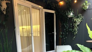 Eine Holzterrasse mit einer Schiebeglastür, die zu einem Duschbereich führt, umgeben von Topfpflanzen und einem weißen Outdoor-Sessel.
