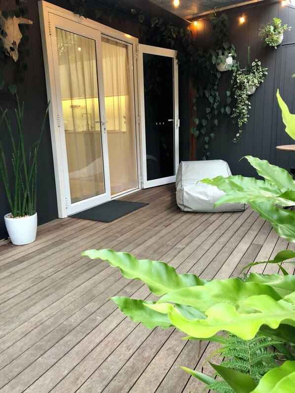 A wooden deck with a sliding glass door leading to a shower area, surrounded by potted plants and a white outdoor chair.