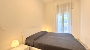 Ein Einzelbett mit grauer Bettdecke, eine Nachttischlampe und ein Fenster mit durchsichtigen Vorhängen.