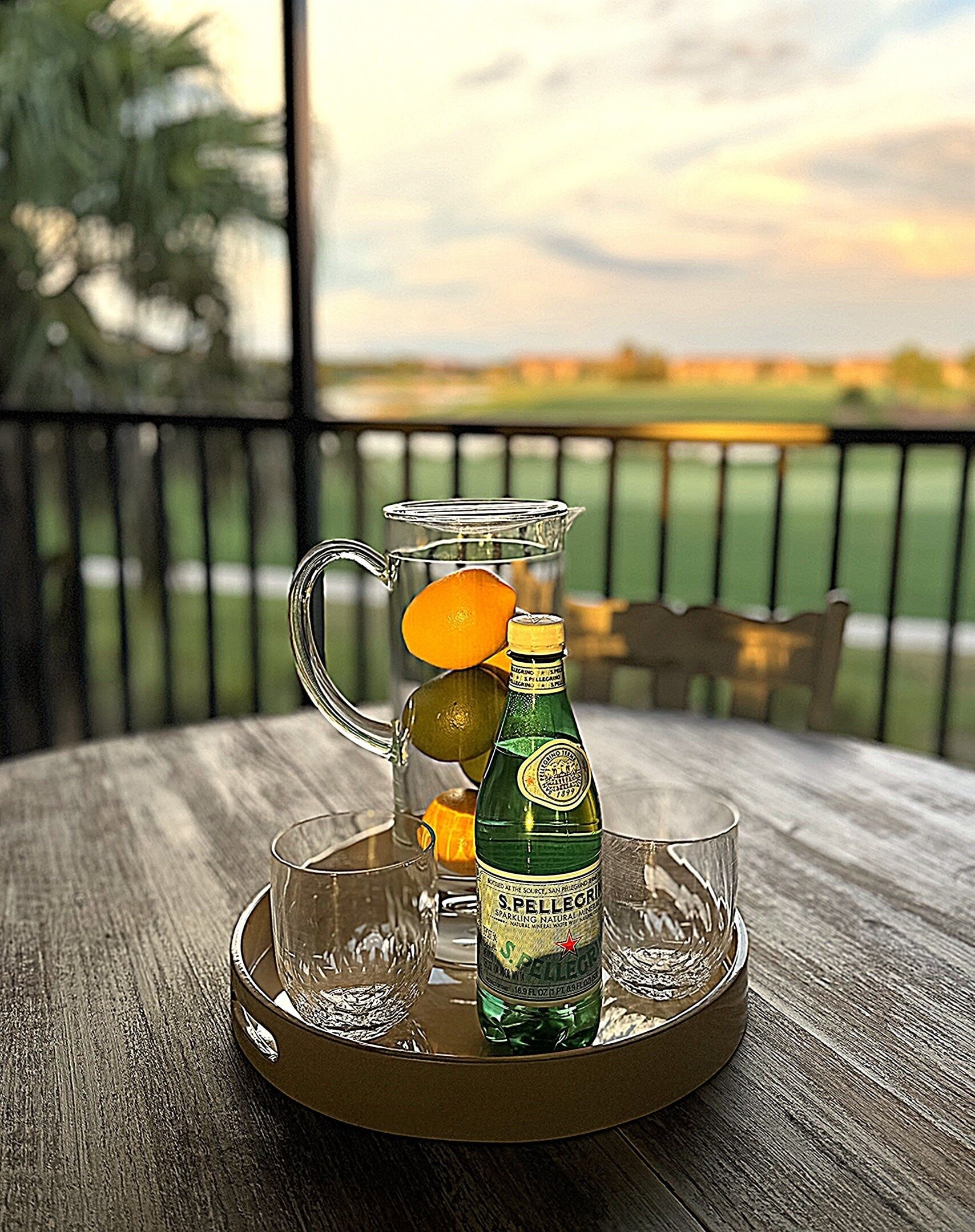 Una jarrón con una botella y vasos sobre una bandeja, colocado en una mesa de madera con vista a un paisaje verde.