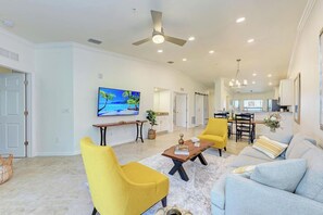 A spacious living room with a ceiling fan, a flat-screen TV, a dining area with a table and chairs, and a kitchen in the background.