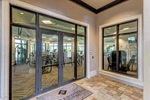 L’entrée d’une salle de sport est dotée de portes vitrées, l’intérieur est équipé de matériel d’exercice et un paillasson avec l’inscription « EWG » est placé devant l’entrée.