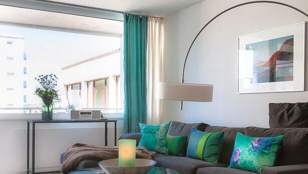 Ein modernes Wohnzimmer mit einem braunen Sofa, farbenfrohen Kissen, einem Glastisch mit Kerze, einer Stehlampe und einem gerahmten Bild an der Wand.