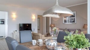 A modern living room with a wooden dining table, grey upholstered chairs, a large metallic pendant light, and a potted plant.