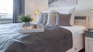 A neatly made bed with a grey and white duvet, a bedside table with a lamp, and a wall-mounted painting.