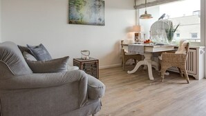 A living room with a grey sofa, a dining table with a chair, a lamp, and a world map painting on the wall. - New Port II - Newport on the beach with south-facing balcony and dune view (Sylt)
